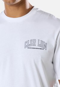 Άνδρας με λευκό μπλουζάκι με μαύρο κείμενο "CLSR LDN ARCHIVE DESIGN DEPARTMENT" στο αριστερό στήθος μπροστά σε απλό φόντο.