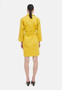 Abito giallo con cintura, maniche lunghe, vita affusolata e lunghezza al ginocchio. Tessuto liscio e stile minimalista.