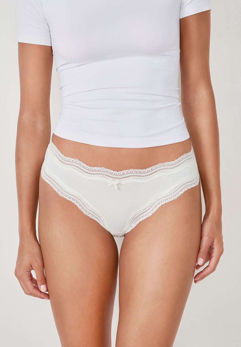 Weiße Unterwäsche mit Spitzenbesatz, aus einem glatten Stoff, mit floralen Details am Rand und einem kleinen Schleifenakzent in der Mitte vorne.