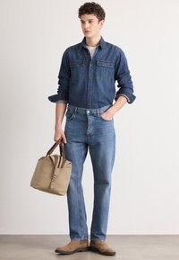 Džinsu krekls ar pogu aizdari, divām krūšu kabatām; gaiši zils džinsu bikses; bēša soma ar brūniem akcentiem; brūnas zamšadas zābaki; neitrāls fons.