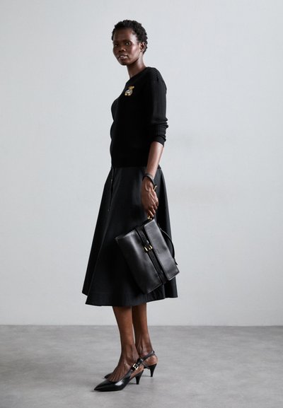 Pull noir à côtes avec une broche décorative, jupe noire en A, sac à main en cuir noir et escarpins à bouts pointus avec brides de cheville.