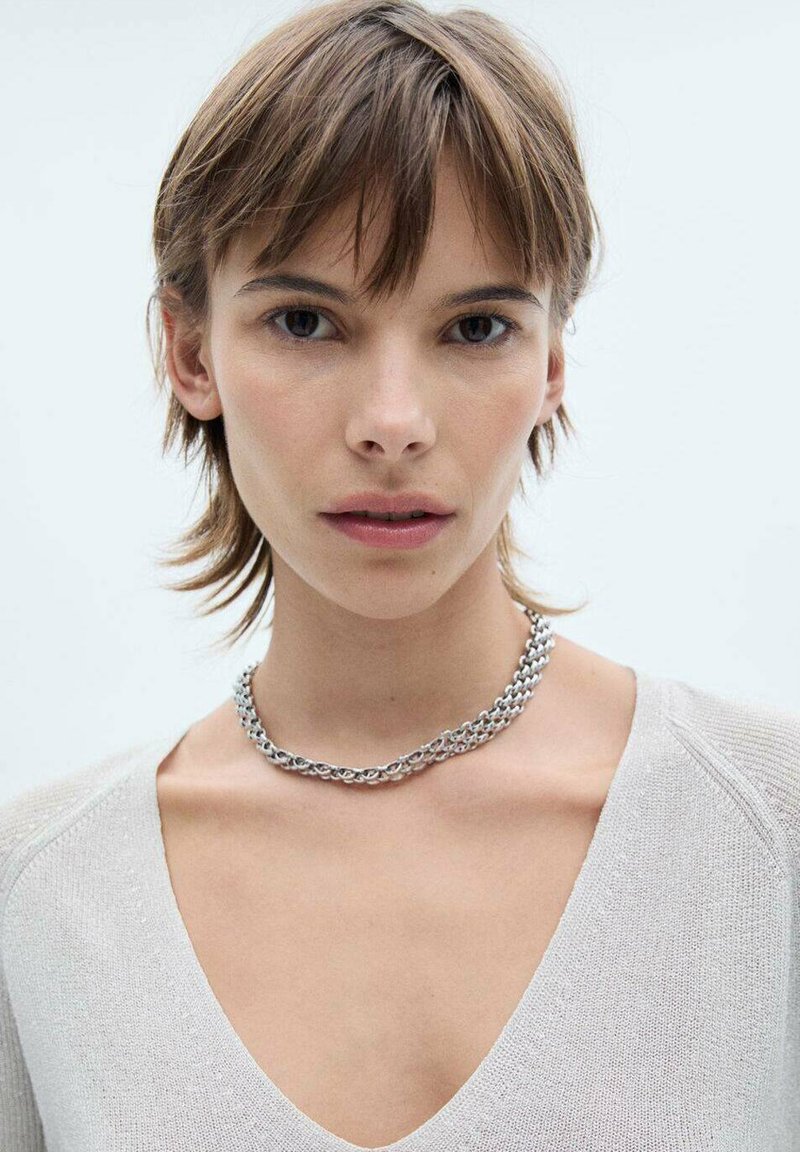 Srebrny naszyjnik łańcuszkowy z połączonymi okrągłymi ogniwami, spoczywający na jasnoszarej, teksturowanej bluzce w kształcie litery V. Neutralne tło.