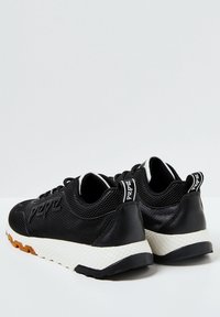 Un paio di sneakers nere in rete con suola bianca testurizzata e suola marrone, viste da dietro e di lato su uno sfondo chiaro.