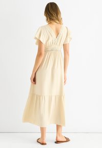 Cream, knee-length dress with short flutter sleeves, lace detailing on the back and waist, and a flowing hemline. Worn with flat sandals.