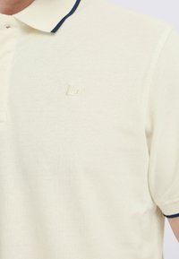 Kremfarget polo-skjorte med teksturert stoff, med en marineblå krage og en subtil brodert logo på brystet.