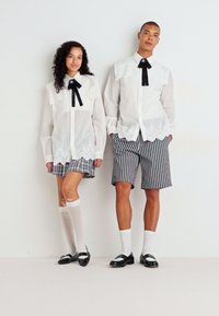 Hvite skjorter med blondesignede krager og svarte bånd, kombinert med stripete shorts. Den kvinnelige modellen har knestrømper; den mannlige modellen har svarte sko.