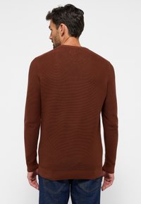 Brauner Langarm-Pullover mit strukturiertem Strickmuster, Rundhalsausschnitt und Rippbündchen, kombiniert mit blauen Jeans im Hintergrund.