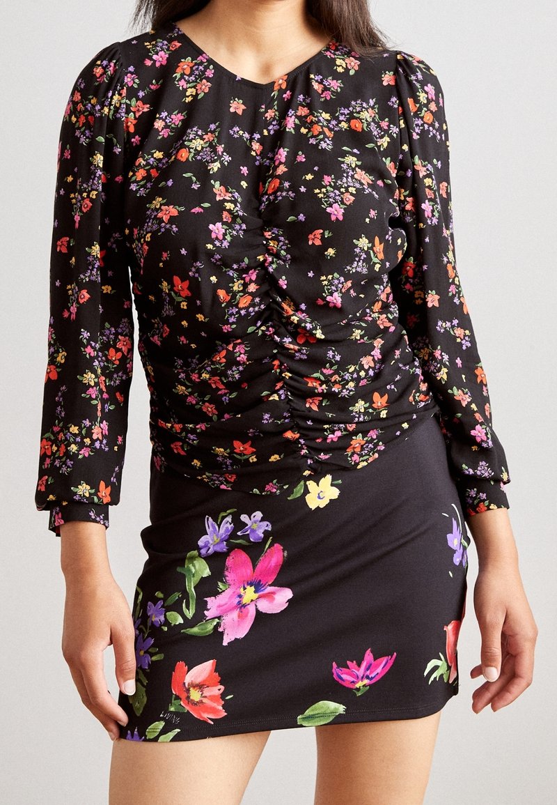 Femme portant une robe noire à fleurs avec des détails froncés sur le haut et des fleurs colorées sur la jupe.