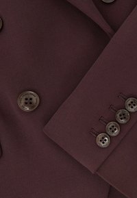 Bordo spalvos pritaikyta švarkas su tekstūruotu paviršiumi, kuriame yra šeši apvalūs, tamsūs mygtukai ir struktūruotas atlapų dizainas.