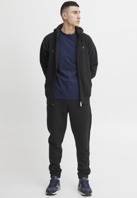 Zwarte zip-up hoodie met zakken, navy crew neck t-shirt, zwarte tapered joggingbroek en navy sport schoenen. Glad materiaal, casual pasvorm.