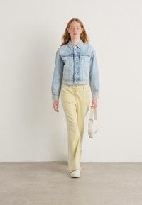 Lichtblauwe spijkerjack met knopen, crèmekleurige blouse, lichtgele broek en witte sneakers, met een kleine lichte tas.