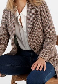 Houndstooth-blazer i beige, burgundy och gröna nyanser; en knapp, notchade slag, tillsammans med en vit skjorta och blå jeans.