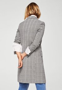 Lungo cappotto a quadri grigio con un design strutturato, rever affilati e maniche bianche arrotolate, abbinato a jeans blu. Presenta una lunghezza a metà schiena e tasche.