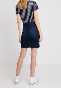 Džínová sukně v tmavě modré barvě, do délky středního stehna, s přiléhavým střihem, zadními kapsami a rozparkem vzadu; v kombinaci s pruhovaným tričkem a bílými teniskami.