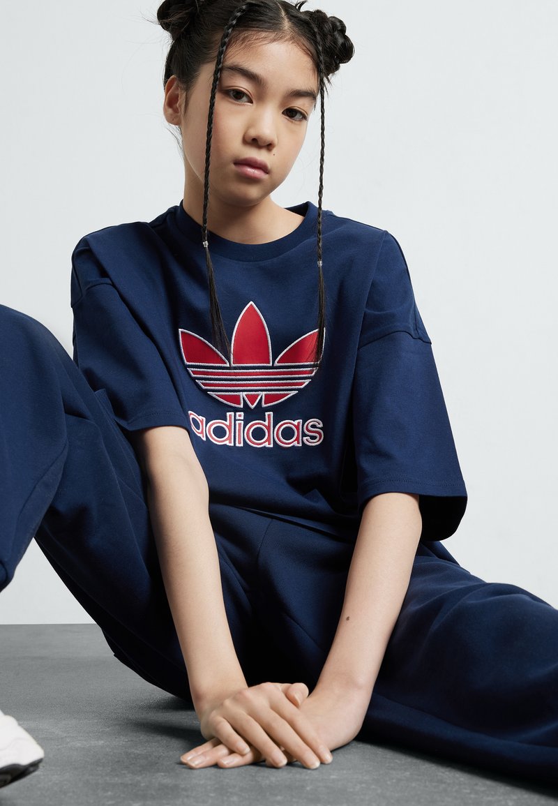 Tumši zila pārspīlēta T-krekls ar sarkano izšūto adidas logo. Tai ir īsas piedurknes un brīvs piegriezums, savienota ar atbilstošām biksēm.