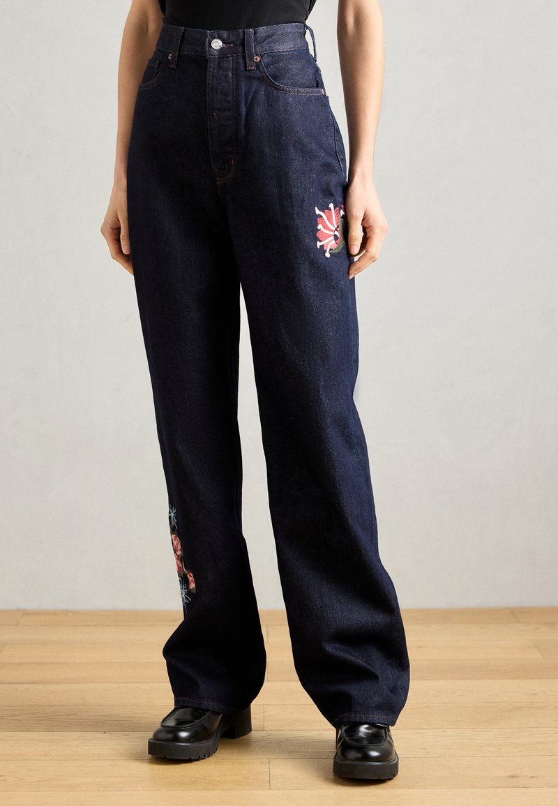 Σκούρο μπλε denim παντελόνι σε άλμα με φαρδύ πόδι, διακοσμημένο με λουλουδάτη κεντήμα στην αριστερή του πλευρά. Σχεδίαση με ψηλή μέση και μεταλλικές λεπτομέρειες σε χαλκό. Μαύρες πλατφόρμες.