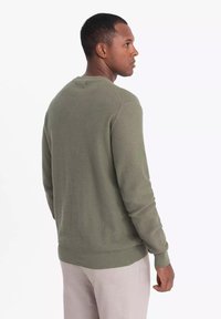 Maglione lavorato a maglia in verde oliva smorzato con un design testurizzato. Presenta un collo a giro e polsini e orlo a coste, indossato sopra un pantalone chiaro.