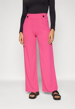 Frau trägt knallpinke, hoch taillierte, weit geschnittene Hose mit schwarzen Knopfdetails, kombiniert mit schwarzem Langarmoberteil und schwarzen Sandalen.