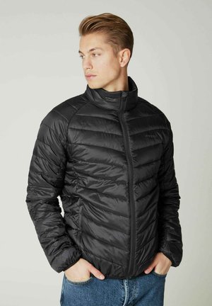 Giacca a vento nera con colletto alto, design trapuntato, chiusura con zip e vestibilità aderente. Indossata sopra un paio di jeans blu, che mettono in risalto una texture liscia e lucida.
