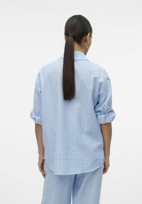 Camicia a righe blu e bianche con maniche arrotolate, vestibilità rilassata e dettaglio della spalla sul retro. Tessuto leggero, design casual e colletto con bottoni.