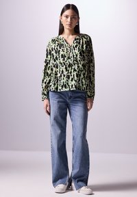 Langärmlige Bluse in grün und schwarz mit Muster und V-Ausschnitt, kombiniert mit hellblauen, weit geschnittenen Jeans und weißen Sneakers. Einfaches Design, leichtes Material.
