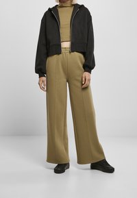 Chaqueta negra con capucha recortada y pantalones anchos de pierna en verde oliva, ambos de tela suave. Los pantalones tienen costuras verticales; combinados con zapatos negros.