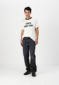White t-shirt with black trim and bold "COACH NEW YORK" text, paired with dark gray jeans featuring a ripped knee and black shoes.