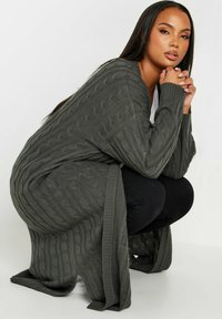 Heathergrön stickad kofta med kabelmönster, lång ärm, öppen fram och ribbad kant. Bärs över en svart topp och byxor.