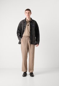 Veste en denim noir oversize avec quincaillerie argentée, polo beige, pantalons beige à taille élastique et chaussures noires. Design minimaliste, sans motifs.