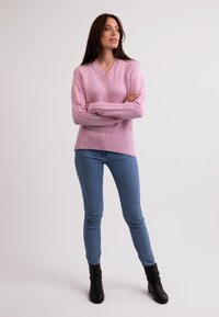 Różowy sweter dziany z dekoltem w kształcie V i ściągaczem, zestawiony z niebieskimi dżinsami i czarnymi botkami za kostkę. Modelka pozująca z rękami skrzyżowanymi.