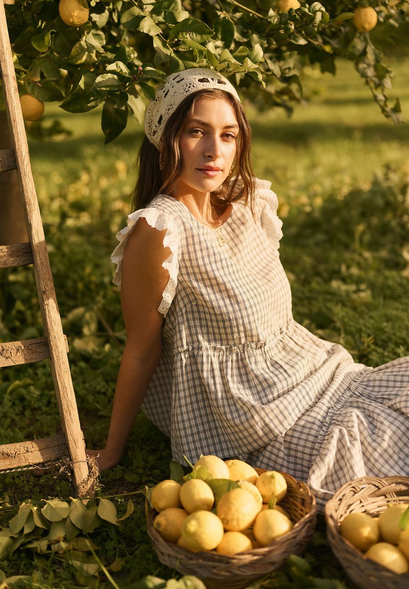 Jeune femme en robe à carreaux et foulard assise près d'une échelle en bois dans un verger de citronniers avec des paniers de citrons sur l'herbe.