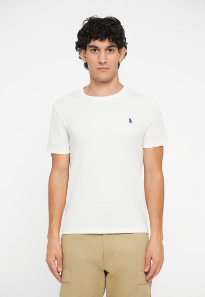 Baltas medvilninis marškinėliai su apvalia iškirpte, trumpomis rankovėmis ir mažuoju mėlynu polo logotipu kairiajame krūtinės šone. Paprastas dizainas, lygus paviršius.