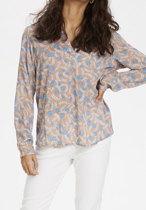 Femme portant un chemisier beige à manches longues avec un motif floral bleu et un pantalon blanc, posant avec une main sur la hanche.