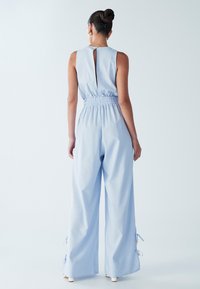 Svetlo modra kombineza z oprijetim pasom in širokimi hlačnicami. Ima odprtino na hrbtu v obliki ključa, elastični trak in dekorativne trakove ob gležnju.