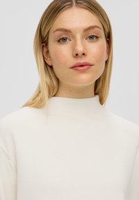 Junge Frau mit blonden Haaren, die einen weißen hochkragen Pullover trägt, direkt nach vorne schaut und einen neutralen Ausdruck auf einem hellen Hintergrund hat.
