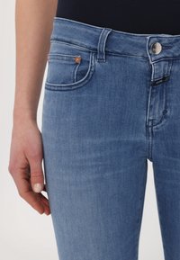 Gros plan sur une personne portant un jean en denim bleu clair avec un bouton et des détails de poche, debout avec la main gauche détendue le long du corps.