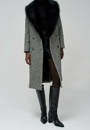 Femme portant un long manteau gris en chevrons avec un col en fourrure noire, une mini-jupe noire, des collants transparents et des bottes noires à talons montant jusqu'aux genoux.