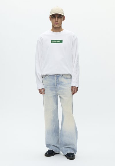 Muž v béžovej čiapke, bielej košeli s dlhým rukávom a nápisom "Mon-Fri:", široké vyblednuté modré rifle a čierne topánky, stojaci pred bielym pozadím.