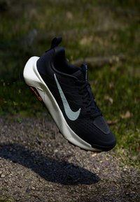 Black athletic shoe with a textured upper, white midsole, and reflective gray swoosh. Features a pull tab and minimal orange branding on the sole.