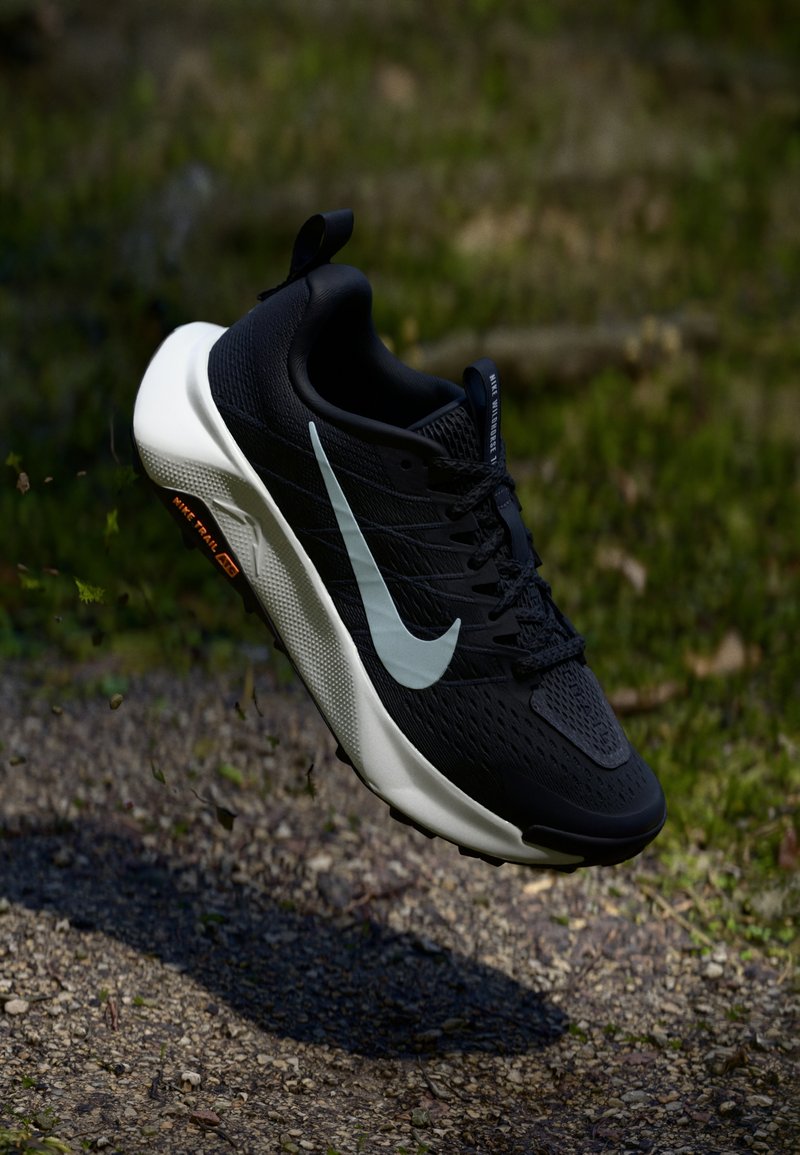 Sort atletik sko med en tekstureret overdel, hvid mellemsål og reflekterende grå swoosh. Har en pull-tab og minimal orange branding på sålen.