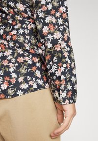 Chemise florale avec un fond noir ornée de fleurs roses, blanches et dorées. Manches longues avec fermeture à bouton au poignet et une texture lisse.