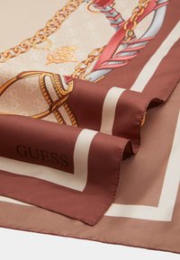 Sciarpa di seta marrone con bordo crema, caratterizzata da un motivo decorativo a catena e nastro. Nome del marchio "GUESS" mostrato in lettere ben visibili.