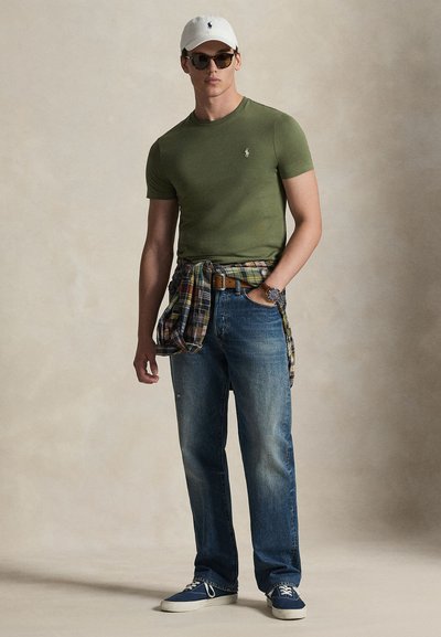 Πράσινο εφαρμοστό μπλουζάκι, μπλε τζιν με ίσια γραμμή, καρό πουκάμισο δεμένο στη μέση, λευκό καπέλο, γυαλιά ηλίου, μπλε αθλητικά παπούτσια. Μινιμαλιστικός σχεδιασμός, casual εμφάνιση.