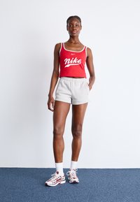 Raudonas nugara su grioveliais marškinėliai su balta Nike logotipu, pilki elastiniai šortai, ir balti kojinės, derinami su raudonais ir baltais sportbačiais. Lygus fonas.