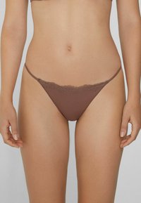 Weiblicher Oberkörper, der braune, spitzenbesetzte, tief sitzende Bikiniunterhosen trägt, vor einem schlichten neutralen Hintergrund.