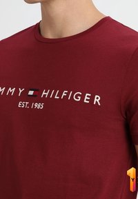 Muž nosící vínovou tričko Tommy Hilfiger s logem a textem "EST. 1985" na prsou.