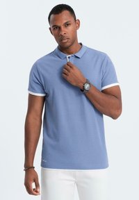 Ljusblå polo med vita detaljer vid kragen och ärmarna, tillverkad av mjukt tyg. Modellen bär den med vita byxor och en klocka.