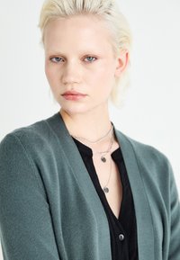 Green knitted cardigan with long sleeves, open front, paired with a black button-up shirt, layered silver necklaces with circular pendants.