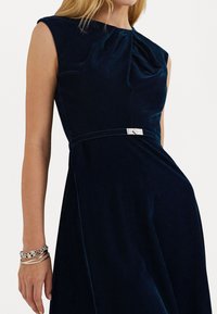 Námornícky zamatový šaty s plisé výstrihom v tvare posádky, bezrukávový dizajn a strieborný kovový detail v oblasti pása. Hladká, textúrovaná látka.