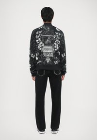 Giacca bomber nera con stampa intricata di fiori e gabbie per uccelli sulla schiena; presenta polsini e orlo a coste; abbinata a jeans neri in denim.